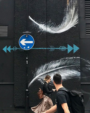 Man and woman walking past a wall with two large feathers painted on it