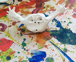 Sculpture of small white creature with arms open wide, on paint-splattered paper